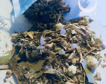 May include: A close-up of a loose leaf tea blend with a variety of herbs, leaves, and flowers. The tea is in a white bag and is spilling out onto a white surface.