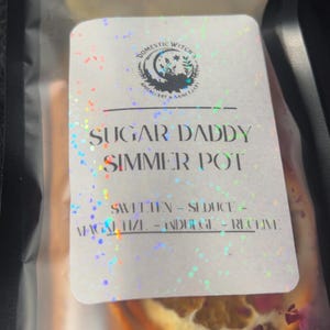 May include: A clear plastic bag containing a simmer pot blend. The label reads "Sugar Daddy Simmer Pot" with the words "Sweeten - Seduce - Magalize - Indulge - Recieve". The bag contains dried orange slices and pink flower petals.