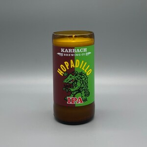 May include: A brown glass candle with a yellow wax top. The candle has a label with a green and red background and a cartoon illustration of a green creature with the text "Hopadilo IPA" and "Karbach Brewing Co."