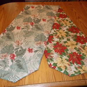 May include: Two decorative table runners. One features a tropical floral pattern with green leaves and red and pink flowers. The other has a Christmas poinsettia design with red and yellow flowers on a white background. Both are on a wooden table.