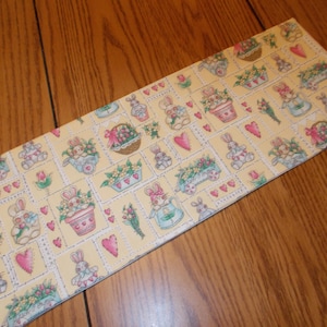 May include: Rectangular Easter-themed fabric with a yellow background and a repeating pattern of bunnies, flowers, and hearts. The design includes bunnies in dresses, flower baskets, and floral arrangements. The fabric is likely used for crafts or home decor.