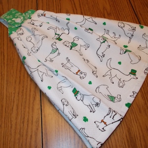 May include: White kitchen towel with a green shamrock pattern and a repeating design of dogs wearing green hats and scarves. The towel has a green loop for hanging.