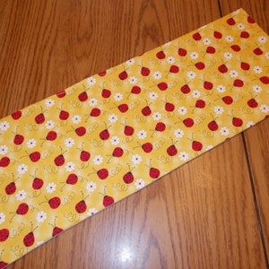 May include: A rectangular fabric item with a yellow background, covered in a repeating pattern of red ladybugs and white daisies. The ladybugs have black spots and antennae. The fabric is likely used for crafting or home decor.