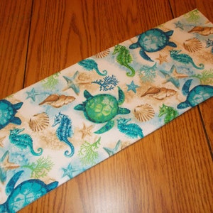 May include: A white table runner with a sea life pattern. The pattern features sea turtles, seahorses, starfish, and seashells in shades of blue, green, and brown.