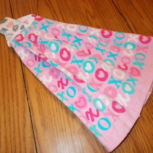 May include: A pink kitchen towel with a pattern of hearts, the letters "x" and "o", and a small floral design. The towel is folded and has a button closure. The towel is made of a soft material.
