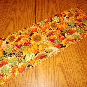 May include: A Thanksgiving table runner with a black background and a colorful pattern of pumpkins, sunflowers, leaves, and gourds.