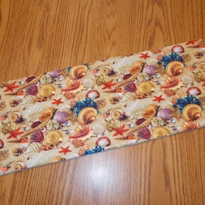 May include: A beige fabric runner with a repeating pattern of seashells, starfish, and other ocean-themed imagery. The fabric has a slightly textured surface.