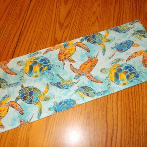 May include: A blue fabric runner with a repeating pattern of colorful sea turtles. The turtles are painted in shades of blue, green, yellow, and orange.