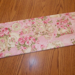 May include: A pink fabric with a floral pattern featuring roses and teacups. The fabric has a light pink background with a watercolor effect. The roses are pink and white, and the teacups are white with gold trim. The fabric has the word "Roses" printed on it.