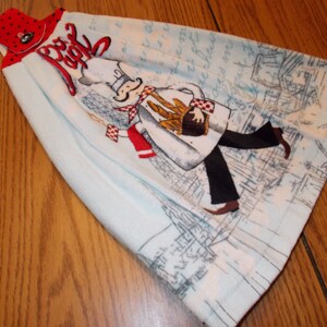 May include: A blue and white kitchen towel with a cartoon chef holding a basket of bread. The towel has a red and white polka dot hanging loop and the text "Ooh La La" in red.