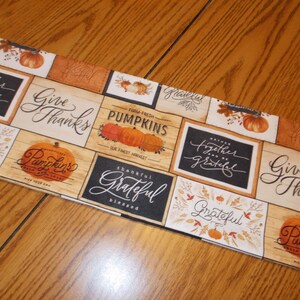 May include: A rectangular fall-themed fabric with a patchwork design. The design features pumpkins, leaves, and phrases like "Give Thanks," "Grateful," and "Pumpkins." The color palette includes orange, brown, and cream.