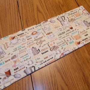 May include: A rectangular coffee-themed placemat with various coffee-related words and images. The placemat features text such as "Coffee," "Espresso," and "Brew," along with illustrations of coffee cups and coffee makers. The background is off-white.