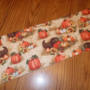 May include: Rectangular fabric with a fall harvest pattern. The design features orange pumpkins, sunflowers, and cornucopias on a beige background. The fabric is likely used for seasonal home decor.