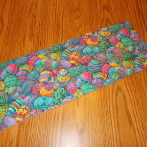 May include: A colorful table runner with a pattern of Easter eggs in shades of blue, green, yellow, pink, and purple. The eggs are arranged in a random pattern and have different designs.