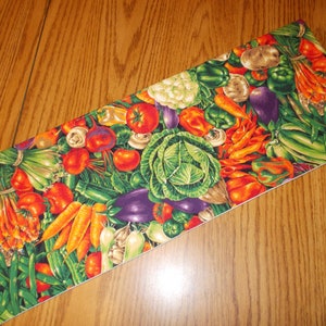 May include: A rectangular fabric piece featuring a vibrant, dense print of assorted fresh vegetables including red tomatoes, orange carrots, green cabbage, purple eggplants, green peppers, and white cauliflower on a dark green background.