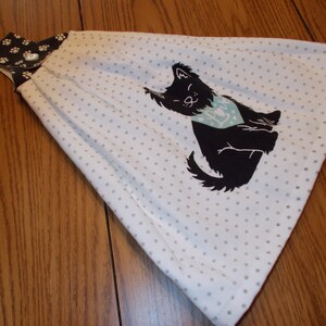 May include: White kitchen towel with a black cat graphic wearing a blue bandana. The towel has a gray polka dot pattern and a dark brown top with paw prints. A button secures the towel.