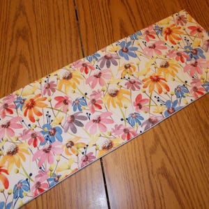 May include: A rectangular floral print table runner featuring a repeating pattern of colorful flowers in shades of pink, orange, yellow, and blue on a cream background. The runner is made of a lightweight fabric and is placed on a wooden table.