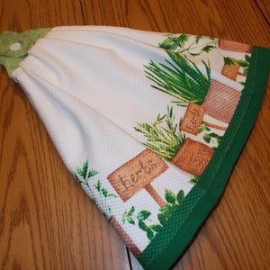 May include: White kitchen towel with a green border. The towel has a printed design of potted herbs with signs that say "herbs" and "seeds".
