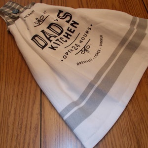 May include: White kitchen towel with gray stripes and the text "DAD'S KITCHEN OPEN 24 HOURS". The towel has a plaid fabric top with a blue button for hanging. The towel is folded on a wooden surface.