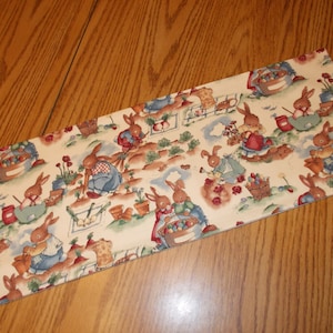May include: Rectangular fabric with a repeating pattern of brown bunnies gardening. The bunnies wear blue and red outfits and tend to flowers and vegetables. Baskets of eggs and gardening tools are also featured.