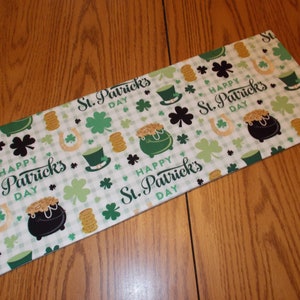 May include: Rectangular St. Patrick's Day fabric with a white and green checkered pattern. The fabric is decorated with green shamrocks, leprechaun hats, horseshoes, pots of gold, and the text "Happy St. Patrick's Day".