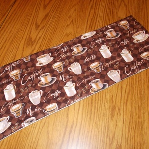 May include: A rectangular coffee-themed table runner. The brown fabric features a checkered pattern with coffee cups, beans, and various coffee drink names like "espresso" and "cappuccino" in white script. The runner has a white border.