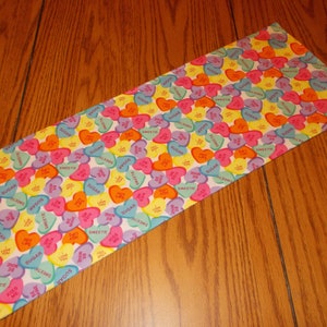 May include: A white fabric runner with a repeating pattern of colorful candy hearts. The hearts have various messages printed on them, including "Sugar", "Sweet", "Kiss Me", and "Love You".