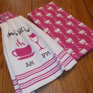 May include: White kitchen towel with pink stripes and a pink coffee cup and wine glass design. The text "How to Tell the Time" is printed on the towel. The towel has a loop for hanging. A pink kitchen towel with a white coffee cup and wine glass pattern is folded on top of the towel.
