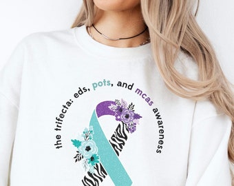 May include: White sweatshirt with a teal and purple ribbon with floral accents. The text around the ribbon reads "the trifecta: eds, pots, and mcas awareness".