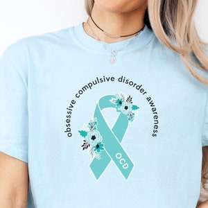 May include: A light blue t-shirt featuring a teal ribbon with a floral design. The ribbon has the text "OCD" and the words "obsessive compulsive disorder awareness" are written around the ribbon.