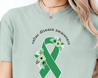Könnte beinhalten: Ein hellgrünes T-Shirt mit dem Text "Celiac Disease Awareness" in einem Kreis um ein grünes Band mit floralen Akzenten.