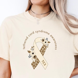 Op de afbeelding: Een ivoorwit T-shirt met een bruin lint met een giraffenprint en bloemmotieven. Het lint heeft de tekst "tethered cord syndrome awareness" en "TCS" erop.