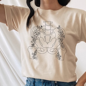 Woman wearing a heather dust colored unisex t-shirt (very light cream), with a black floral pelvis anatomy design on it. The design is large in the center of the shirt. She has black hair pulled up, and jeans on.