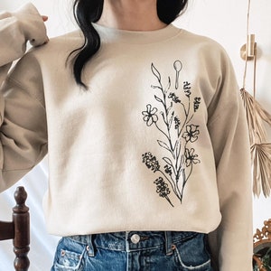 May include: A beige sweatshirt featuring a black line drawing of a floral bouquet on the front. The sweatshirt is being worn by a person wearing denim shorts.
