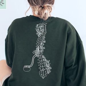 Puede incluir: Una sudadera verde oscuro con un contorno blanco de una columna vertebral con flores que crecen de ella. La columna vertebral está conectada a una pequeña bomba.