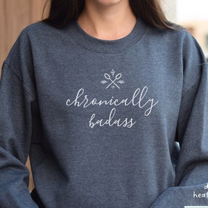 Op de afbeelding: Een donker grijs sweatshirt met de tekst "chronically badass" in witte letters. Boven de tekst staat een kleine afbeelding van gekruiste scharen met een hart.