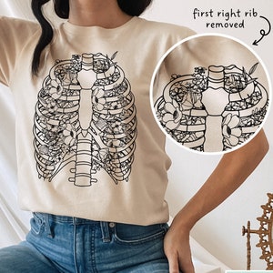 May include: A beige t-shirt with a black line drawing of a rib cage filled with flowers. The text "first right rib removed" is written above the image.