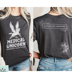 May include: A dark grey t-shirt with a white unicorn graphic and the text "The Medical Unicorn Misunderstood & Always a Mystery". The back of the shirt has the text "Nutcracker Syndrome" in a white rectangle with floral designs.