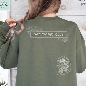 May include: A military green sweatshirt with the text "ONE KIDNEY CLUB" in white lettering. The sweatshirt features a white outline of a kidney with floral details.