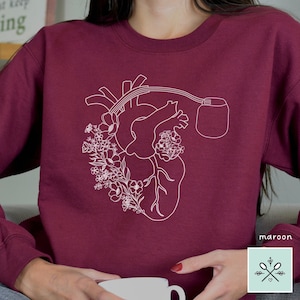 May include: A maroon sweatshirt with a white outline of a heart with flowers and a medical device attached to it. The text "maroon" is visible in the bottom right corner of the image.