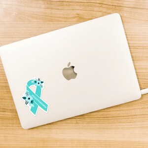 Op de afbeelding: Een turquoise lint met witte bloemen en het woord "DYSAUTONOMIA" op een witte laptop.