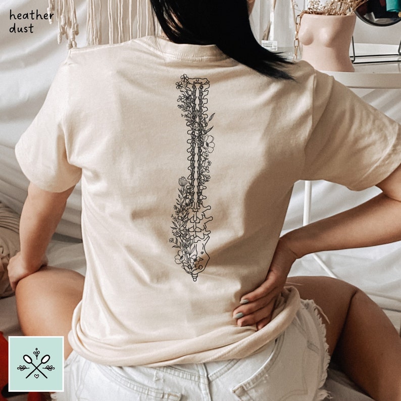 Woman sitting with her back facing the camera wearing a heather dust colored t-shirt. The back of the shirt has a black floral spinal fusion anatomy design on it, with the spine straight down the center of the shirt. She is wearing white jean shorts.