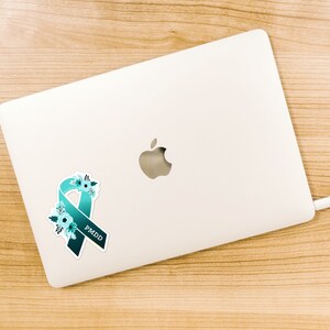 May include: A teal blue ribbon with white flowers and the text "PMDD" on a white laptop.
