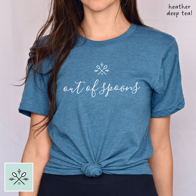 Woman wearing a heather deep teal colored unisex t-shirt. The shirt says out of spoons in pretty white script writing centered across the chest. There is a white crossed spoons spoonie logo symbol above the text, for decoration.