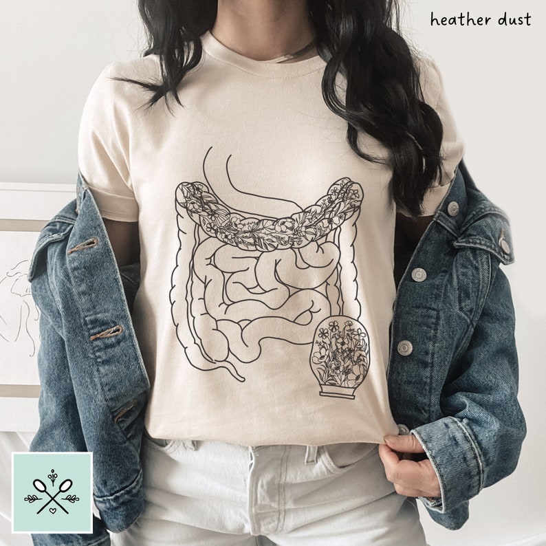 May include: A beige t-shirt with a black line drawing of the human digestive system with flowers inside. The text "heather dust" is in the top right corner of the image.
