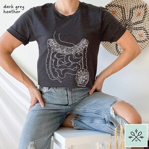 May include: A dark grey heather t-shirt with a white outline of the human digestive system and a floral design. The shirt is being worn by a person wearing blue jeans.