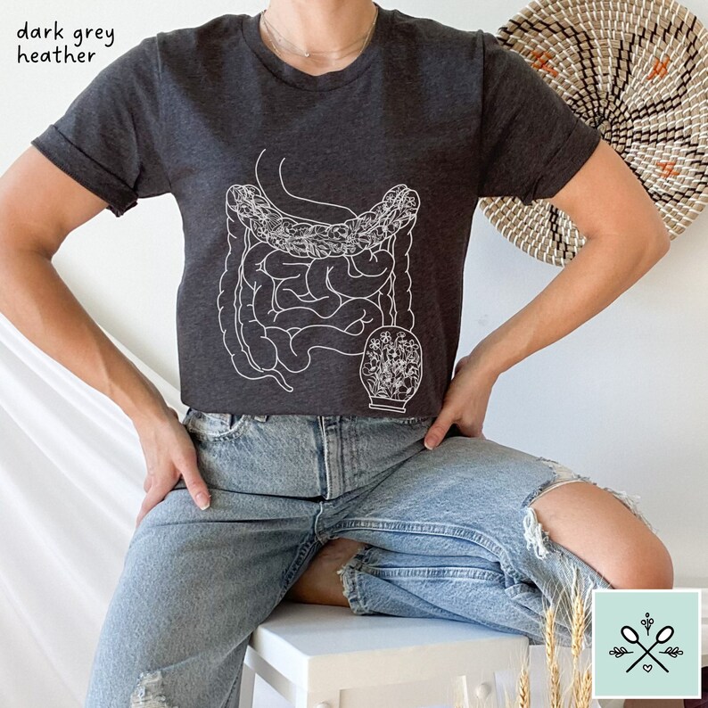 May include: A dark grey heather t-shirt with a white outline of the human digestive system and a floral design. The shirt is being worn by a person wearing blue jeans.