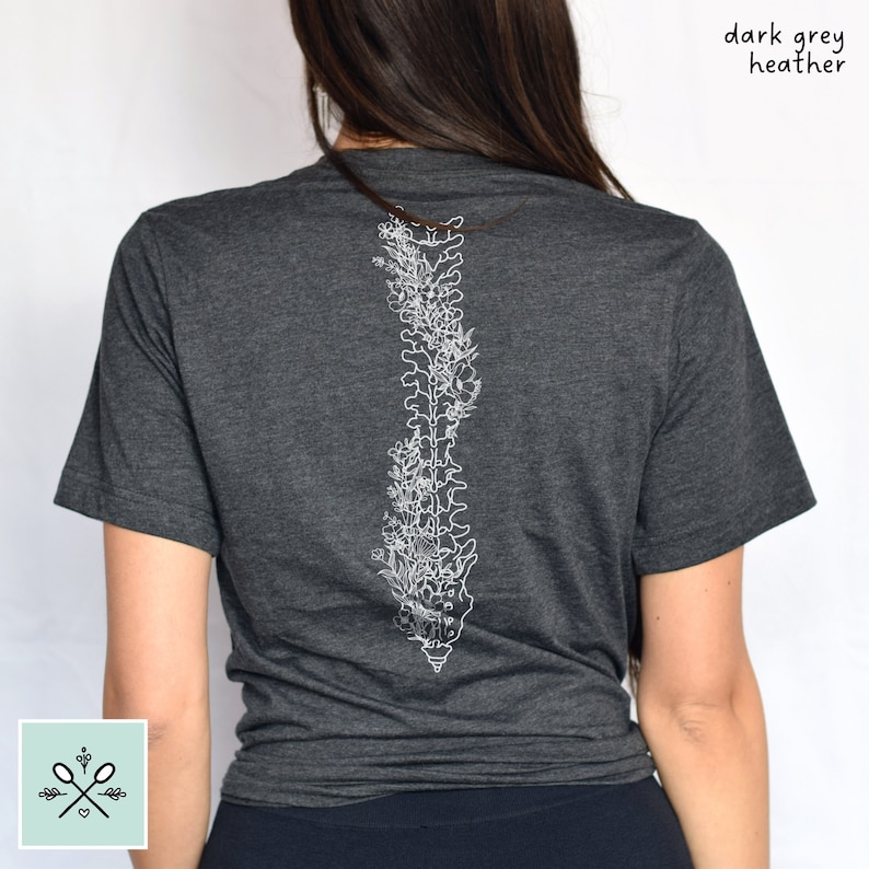 Woman wearing a dark grey heather unisex t-shirt, with her back to the camera. There is a white floral spine outline design going down the center of her back on the t-shirt. The artistic design starts at the top of the shirt and goes to her mid-back.