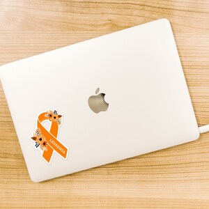 May include: Orange ribbon with floral details and the word "Leukemia" on a white laptop.