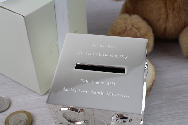 May include: Silver-colored piggy bank shaped like alphabet blocks with a slot for coins. The bank is engraved with the text "Willow Grace", "On Your Christening Day", "29th August 2021", and "All My Love Auntie Marie xxx".